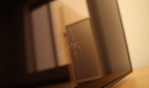 Close-up goresan halus berbentuk silang pada permukaan layar perangkat dengan latar belakang ruangan yang tampak buram.